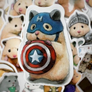 May include: A sticker of a hamster wearing a blue and red Captain America costume, holding a red, white, and blue shield with a white star in the center. The hamster is surrounded by other hamster stickers.