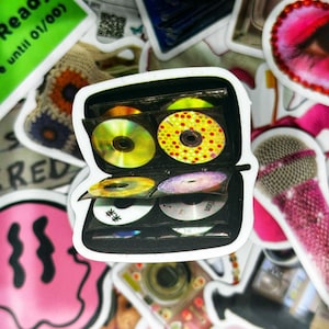 May include: A black CD storage case with a clear plastic cover. The case is open and filled with colourful CDs. The case is a popular way to store and organise CDs.