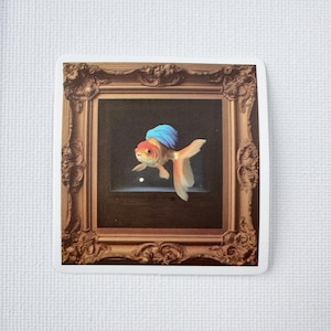 May include: A sticker depicting a goldfish wearing a blue turban, framed within an elaborate, gold-coloured picture frame. The goldfish has orange and white fins and a red head. The background is dark.