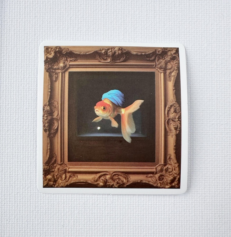 May include: A sticker depicting a goldfish wearing a blue turban, framed within an elaborate, gold-coloured picture frame. The goldfish has orange and white fins and a red head. The background is dark.