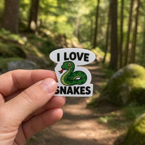 May include: A white sticker featuring the words "I LOVE SNAKES" and a cartoon green snake with a red tongue. The sticker is held against a blurred forest backdrop.