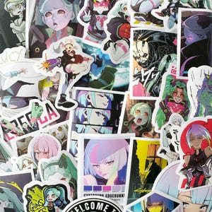 Peut inclure: Une collection d'autocollants de style anime colorés mettant en scène des personnages aux cheveux et aux vêtements vibrants. Les autocollants ont un thème cyberpunk et incluent du texte comme "Cyberpunk Edgerunners (2022)" et "Bienvenue à".