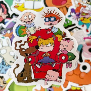 May include: A sticker featuring the Rugrats characters, including Tommy Pickles, Chuckie Finster, Phil and Lil DeVille, and Angelica Pickles. The characters are sitting on a red armchair with a brown dog standing next to them. The sticker is surrounded by other cartoon stickers.