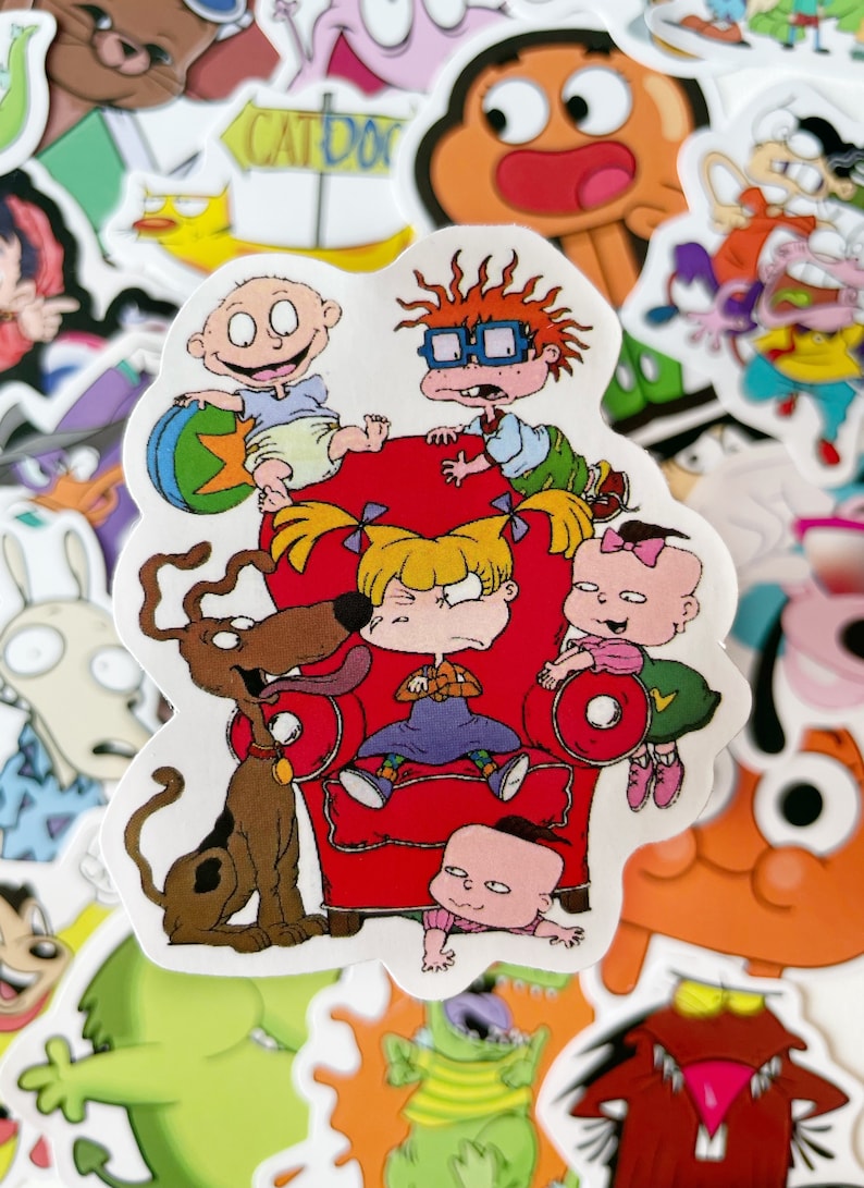 May include: A sticker featuring the Rugrats characters, including Tommy Pickles, Chuckie Finster, Phil and Lil DeVille, and Angelica Pickles. The characters are sitting on a red armchair with a brown dog standing next to them. The sticker is surrounded by other cartoon stickers.