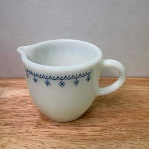 May include: A white, vintage creamer with a blue snowflake design around the rim. The creamer has a handle and a spout for easy pouring. It is made of opaque glass and sits on a wooden surface.