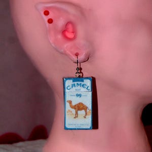 Op de afbeelding: Een opvallende oorbel met een miniatuur Camel Blue sigarettenpakje. Het pakje is blauw en wit met een kameelafbeelding. De oorbel hangt aan een zilverkleurige haak. De oorbel wordt aan een oor getoond.