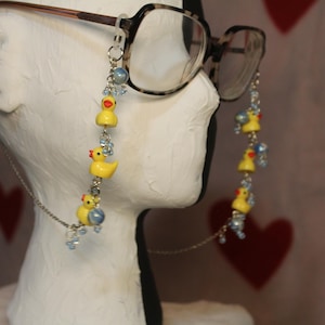 May include: Eyeglass chain with yellow duck charms, red beaks, and blue bead accents. The chain is attached to tortoiseshell eyeglasses. The background is light pink with red heart shapes.