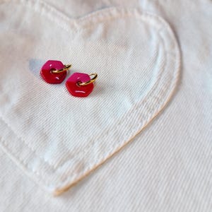 May include: A pair of gold hoop earrings with pink and red hexagonal charms. The earrings are on a white fabric background.