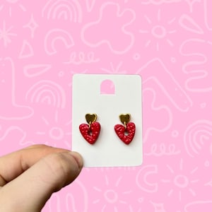 May include: A pair of red heart-shaped earrings with gold accents. The earrings are displayed on a white card.