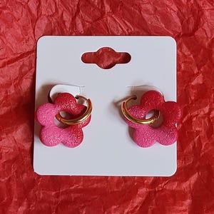 May include: A pair of flower-shaped earrings in shades of pink and red, attached to gold-coloured hoops. The earrings are presented on a white card, set against a red backdrop.