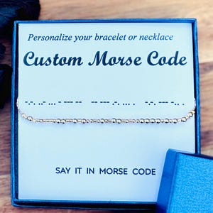 May include: A gold-colored Morse code bracelet displayed in a blue gift box. The box contains a card that reads "Personalize your bracelet or necklace" and "Custom Morse Code". The text "Say it in Morse Code" is also visible.