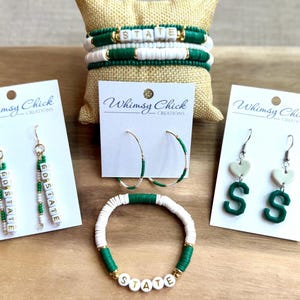 May include: Jewelry set featuring bracelets and earrings in green, white, and gold. The bracelets spell out "STATE", and the earrings have "GO STATE" and "S" designs. The brand "Whimsy Chick Creations" is visible on the cards.