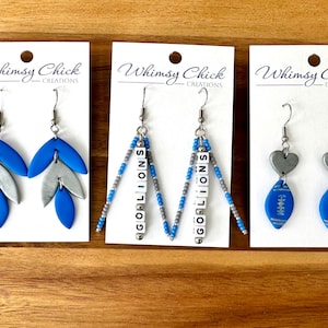 May include: Three pairs of earrings on white cards. One pair has blue and silver leaf shapes. Another has beads spelling "GO LIONS". The last pair has silver hearts and blue football shapes. The cards have the text "Whimsy Chick Creations".
