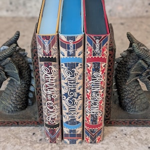 Puede incluir: Tres libros de tapa dura con cubiertas de patrones rojos, azules y blancos. Los t&iacute;tulos son "Shadow and Bone", "Siege and Storm" y "Ruin and Rising" de Leigh Bardugo.