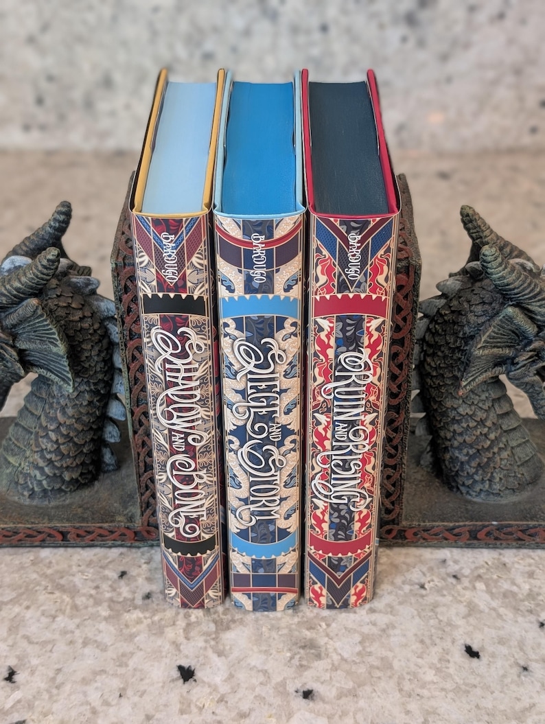 Puede incluir: Tres libros de tapa dura con cubiertas de patrones rojos, azules y blancos. Los t&iacute;tulos son "Shadow and Bone", "Siege and Storm" y "Ruin and Rising" de Leigh Bardugo.