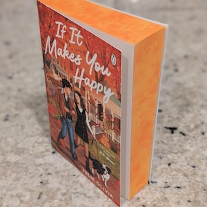 Puede incluir: Un libro de bolsillo titulado "If It Makes You Happy" de Julie Olivia. La portada presenta a una pareja paseando a un perro, sobre un fondo de follaje oto&ntilde;al y una casa. El libro tiene lomo y bordes naranjas.