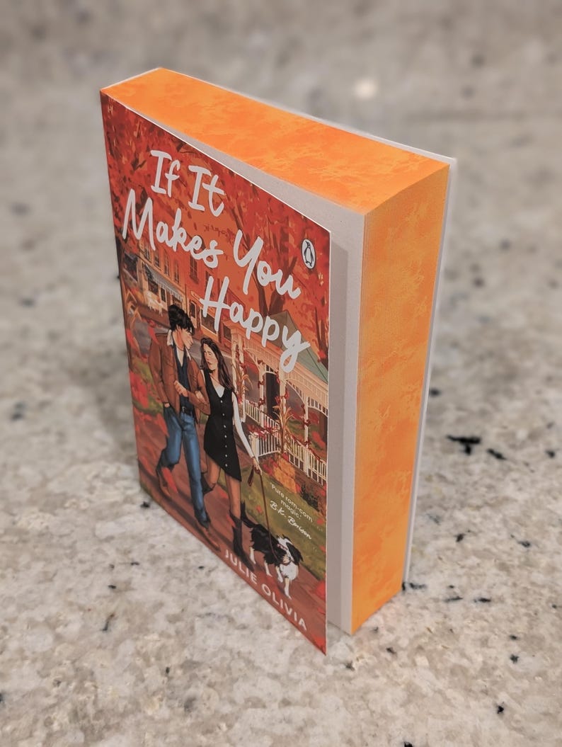 Puede incluir: Un libro de bolsillo titulado "If It Makes You Happy" de Julie Olivia. La portada presenta a una pareja paseando a un perro, sobre un fondo de follaje oto&ntilde;al y una casa. El libro tiene lomo y bordes naranjas.