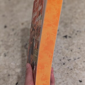 Puede incluir: Un libro titulado "If It Makes You Happy" con lomo naranja y portada con un grupo de personas. El libro est&aacute; sostenido por una persona.