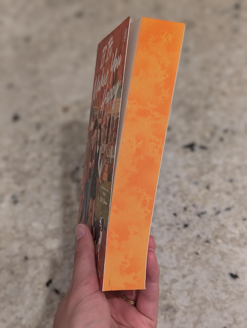 Puede incluir: Un libro titulado "If It Makes You Happy" con lomo naranja y portada con un grupo de personas. El libro est&aacute; sostenido por una persona.