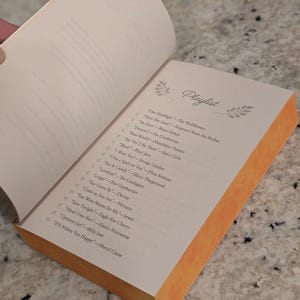 Puede incluir: Un libro abierto con una lista de reproducci&oacute;n titulada "Playlist" en la p&aacute;gina derecha. La p&aacute;gina tiene una lista de canciones y artistas. El libro tiene un borde naranja y est&aacute; sobre una superficie gris.