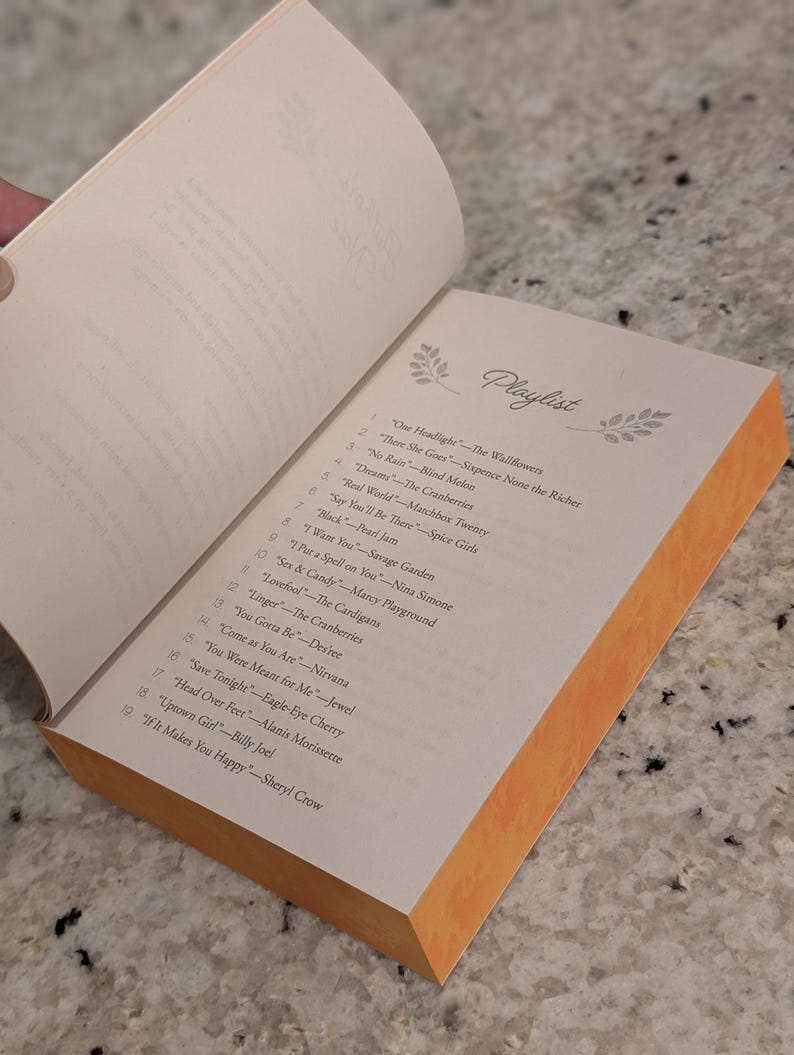 Puede incluir: Un libro abierto con una lista de reproducci&oacute;n titulada "Playlist" en la p&aacute;gina derecha. La p&aacute;gina tiene una lista de canciones y artistas. El libro tiene un borde naranja y est&aacute; sobre una superficie gris.