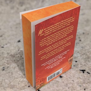 Puede incluir: Un libro de bolsillo con una cubierta naranja y roja. El lomo del libro es visible, mostrando el t&iacute;tulo y el nombre del autor. La contraportada presenta texto y una peque&ntilde;a imagen de un edificio. El libro est&aacute; ligeramente inclinado, revelando las p&aacute;ginas blancas.