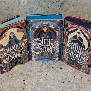 Puede incluir: Tres libros de tapa dura con cubiertas coloridas que presentan ilustraciones de personajes y texto. Los t&iacute;tulos son "Shadow and Bone", "Siege and Storm" y "Ruin and Rising".