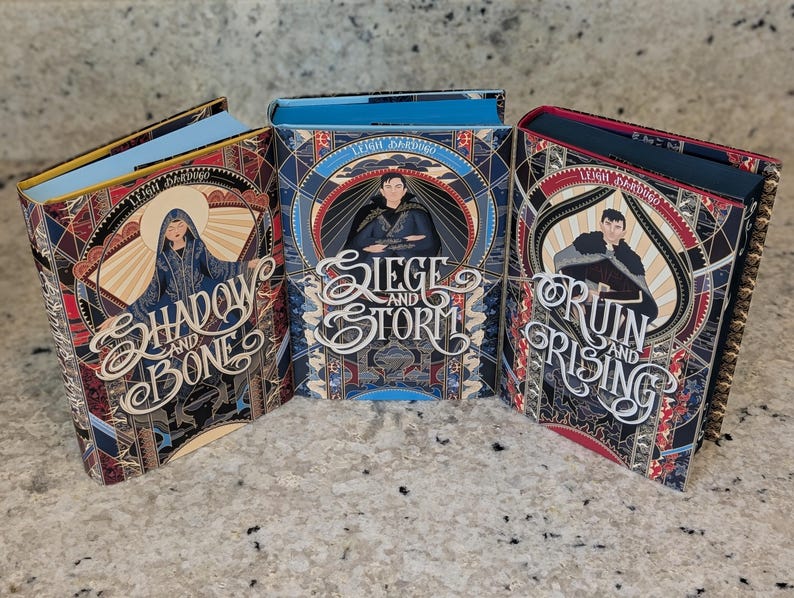 Puede incluir: Tres libros de tapa dura con cubiertas coloridas que presentan ilustraciones de personajes y texto. Los t&iacute;tulos son "Shadow and Bone", "Siege and Storm" y "Ruin and Rising".