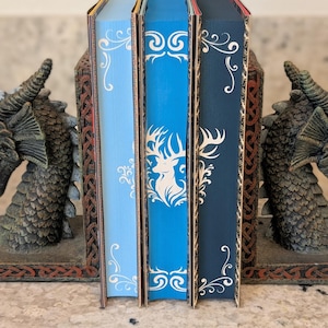 Puede incluir: Tres sujetalibros decorativos con un dise&ntilde;o de drag&oacute;n. Los sujetalibros est&aacute;n hechos de un material marr&oacute;n oscuro con detalles dorados. Los sujetalibros sostienen tres libros con cubiertas azules, azul claro y azul oscuro. Los libros tienen un dise&ntilde;o blanco en la portada.