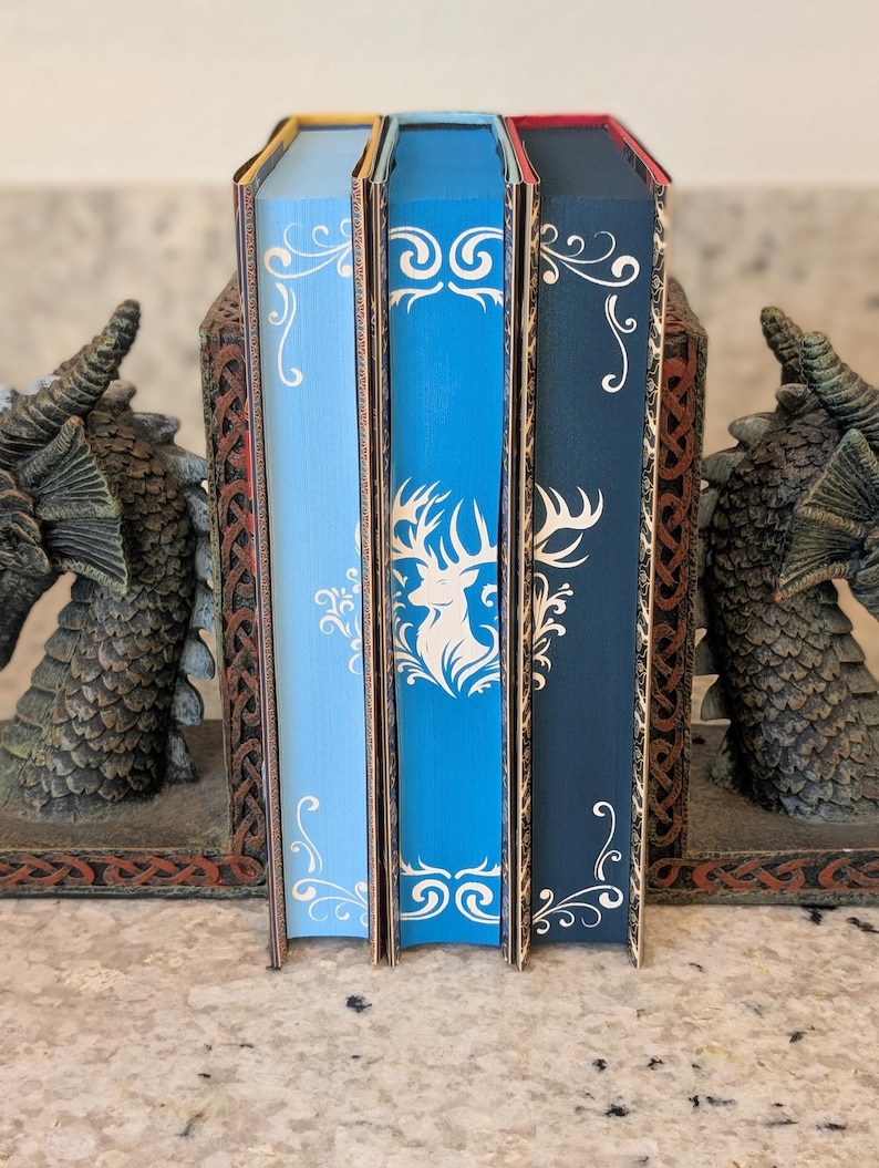 Puede incluir: Tres sujetalibros decorativos con un dise&ntilde;o de drag&oacute;n. Los sujetalibros est&aacute;n hechos de un material marr&oacute;n oscuro con detalles dorados. Los sujetalibros sostienen tres libros con cubiertas azules, azul claro y azul oscuro. Los libros tienen un dise&ntilde;o blanco en la portada.