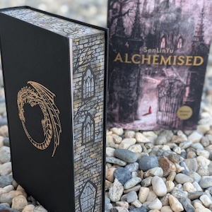 May include: A black book with a gold dragon design and the title "ALCHEMISED" on the cover. The spine features a brick and window design. The book is set on a bed of small, gray stones.