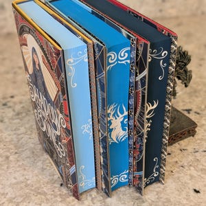 Puede incluir: Cinco libros de tapa dura con diferentes colores de lomo y dise&ntilde;os intrincados. Los lomos son rojo, amarillo, azul, azul y negro. Los libros tienen detalles dorados y patrones decorativos.