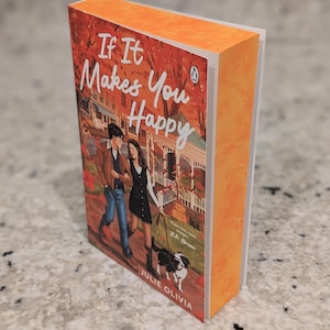 Puede incluir: Un libro de bolsillo titulado "If It Makes You Happy" de Julie Olivia. La portada muestra a una pareja caminando con un perro. El libro tiene el lomo y los bordes naranjas, con el logo de Penguin.