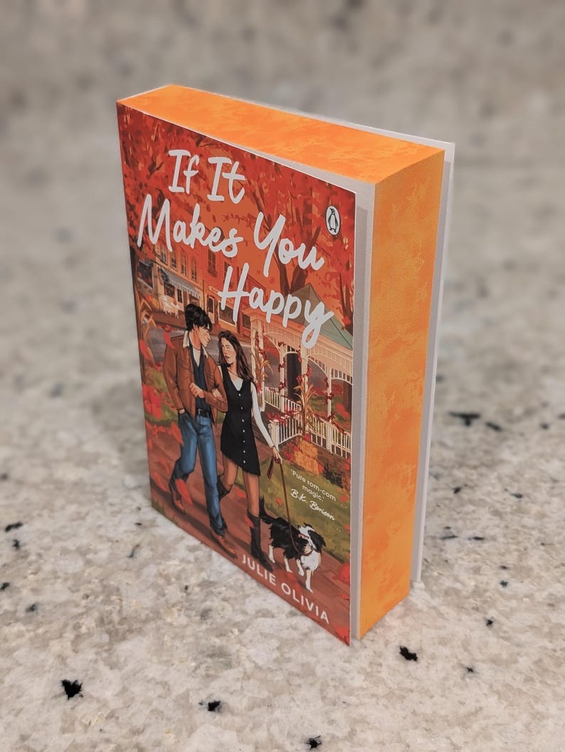 Puede incluir: Un libro de bolsillo titulado "If It Makes You Happy" de Julie Olivia. La portada muestra a una pareja caminando con un perro. El libro tiene el lomo y los bordes naranjas, con el logo de Penguin.