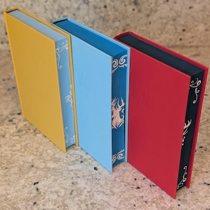 Puede incluir: Tres libros de tapa dura con cubiertas de diferentes colores. El libro de la izquierda es amarillo, el libro del medio es azul claro y el libro de la derecha es rojo.