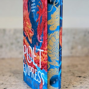 Op de afbeelding: Een hardcover boek getiteld "The Poet Empress" van Shen Tao. De omslag toont een levendig rood drakenontwerp tegen een blauwe achtergrond. Gouden draken- en sterdetails staan op de rug.
