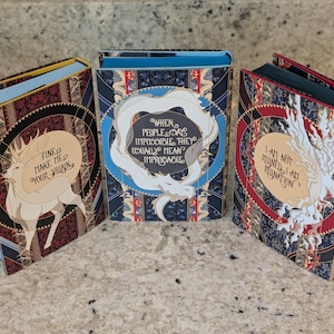 Puede incluir: Tres libros de tapa dura con cubiertas coloridas que presentan criaturas m&iacute;ticas blancas y texto. El primer libro tiene una cubierta azul y roja con el texto "Fine make me your villain". El segundo libro tiene una cubierta azul y dorada con el texto "When people say impossible they usually mean improbable". El tercer libro tiene una cubierta roja y dorada con el texto "I am not ruined I am a ruin".