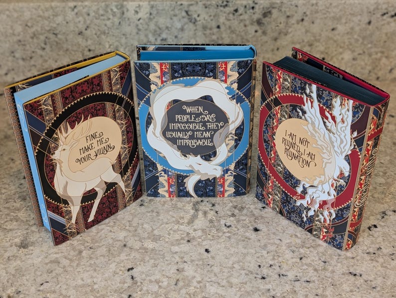 Puede incluir: Tres libros de tapa dura con cubiertas coloridas que presentan criaturas m&iacute;ticas blancas y texto. El primer libro tiene una cubierta azul y roja con el texto "Fine make me your villain". El segundo libro tiene una cubierta azul y dorada con el texto "When people say impossible they usually mean improbable". El tercer libro tiene una cubierta roja y dorada con el texto "I am not ruined I am a ruin".