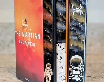 May include: A hardcover copy of The Martian by Andy Weir with a red and orange cover featuring an astronaut. The book's page edges are hand-painted with a black space scene, white stars, a moon surface, and two astronaut figures in white.