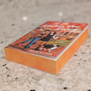 Puede incluir: Un libro de bolsillo titulado "Makes You Happy" de Julie Olivia. La portada presenta una ilustraci&oacute;n de una pareja paseando a un perro. El libro tiene una cubierta naranja y est&aacute; sobre una superficie moteada.