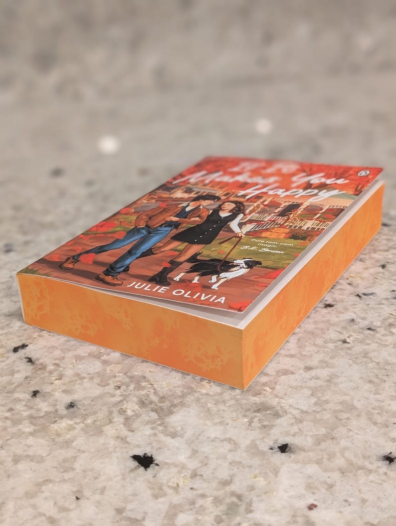 Puede incluir: Un libro de bolsillo titulado "Makes You Happy" de Julie Olivia. La portada presenta una ilustraci&oacute;n de una pareja paseando a un perro. El libro tiene una cubierta naranja y est&aacute; sobre una superficie moteada.