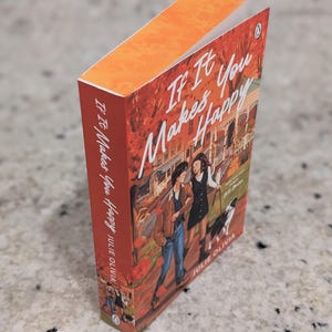 Puede incluir: Un libro de bolsillo titulado "If It Makes You Happy" de Julie Olivia. La portada presenta una ilustraci&oacute;n de dos personas caminando con un perro. El libro tiene un lomo naranja y una cubierta roja y naranja.