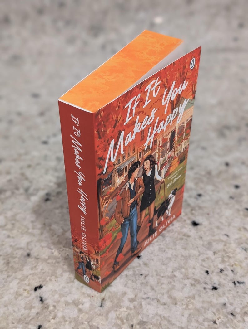 Puede incluir: Un libro de bolsillo titulado "If It Makes You Happy" de Julie Olivia. La portada presenta una ilustraci&oacute;n de dos personas caminando con un perro. El libro tiene un lomo naranja y una cubierta roja y naranja.