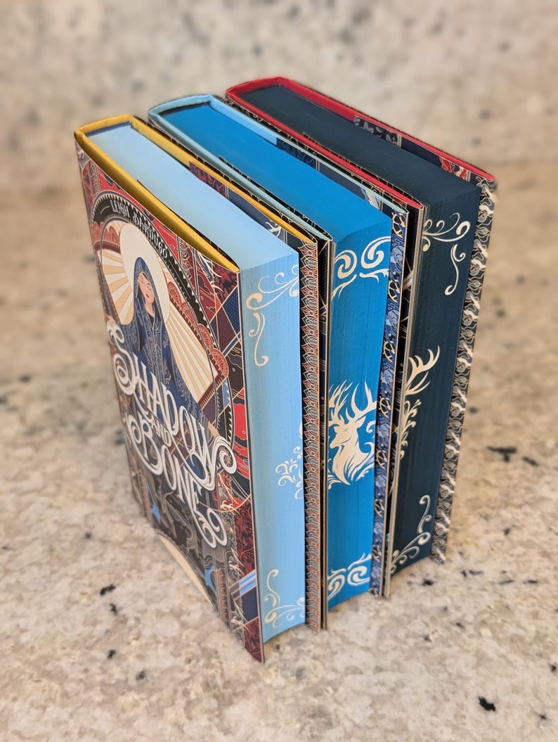 Puede incluir: Cuatro libros de tapa dura con lomos azules, verde azulado y rojos. El libro de la izquierda tiene una portada colorida con el t&iacute;tulo "Shadow and Bone" y el nombre de la autora Leigh Bardugo. Los otros libros tienen un dise&ntilde;o m&aacute;s simple con un patr&oacute;n de ciervo y una ola.
