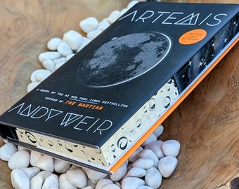 Puede incluir: Un libro de tapa dura titulado Artemis de Andy Weir con una cubierta negra que presenta una ilustración de la luna en blanco, detalles en naranja y una pegatina de New York Times Bestseller. Los bordes tienen un patrón de fases lunares.