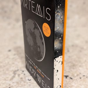May include: A hardcover book titled "Artemis" by Andy Weir. The cover is black with a gray illustration of the moon and white text. The spine is orange with a starry design. A New York Times Bestseller sticker is on the cover.
