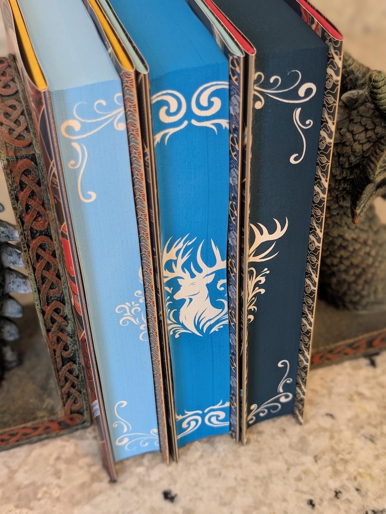 Puede incluir: Cuatro libros de tapa dura con lomos decorativos. Los lomos son azules, azul claro, azul oscuro y rojo. Los libros tienen dise&ntilde;os decorativos blancos en los lomos. Los libros est&aacute;n dispuestos en una fila de izquierda a derecha.