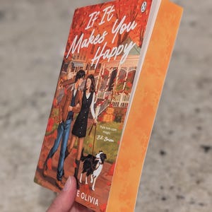 Puede incluir: Un libro de bolsillo titulado "If It Makes You Happy" de B.K. Borison. La portada presenta a una pareja paseando a un perro, con un fondo oto&ntilde;al. El libro tiene el lomo naranja y se sostiene en &aacute;ngulo.