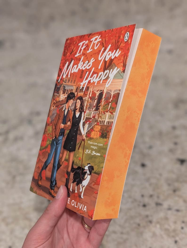 Puede incluir: Un libro de bolsillo titulado "If It Makes You Happy" de B.K. Borison. La portada presenta a una pareja paseando a un perro, con un fondo oto&ntilde;al. El libro tiene el lomo naranja y se sostiene en &aacute;ngulo.