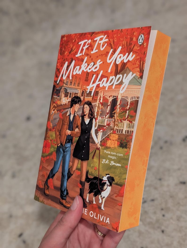 Puede incluir: Un libro de bolsillo titulado "If It Makes You Happy" con una ilustraci&oacute;n de portada de una pareja caminando con un perro. La portada tiene una combinaci&oacute;n de colores naranja y rojo con el nombre del autor, Jamie Olivia, en la parte inferior.